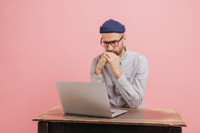 Horizontal shot of concentrated male worker reads statistics attentively, focused in laptop computer, learns something online, wears spectacles, hat and white shirt, isolated over pink wall.